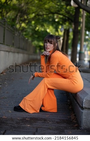 Portrait of a beautiful caucasian woman in her 20s wearing an orange outfit. She is sitting and posing. She has brown hair and is wearing glasses. 