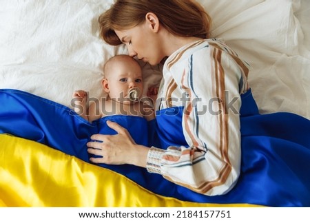 Close-up portrait of a mother kissing her newborn baby sleeping on the bed at home covered with the Blue and yellow flag of Ukraine. The baby sleeps with the safety and protection of the mother