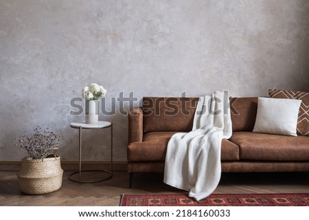 Loft design of living room interior with brown eco leather couch, knitted blanket, cushions, side table with blooming flowers in ceramic vase and wicker basket on the floor Royalty-Free Stock Photo #2184160033