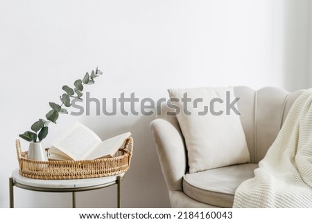 Wicker tray and open book close to eucalyptus branch in ceramic vase on side table. Bohemian design of living room with beige soft chair with cushion and white plaid Royalty-Free Stock Photo #2184160043