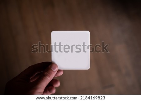 Hand holding a blank white square paper card with rounded corners. Business card, magnet, bumper sticker mock-up. Wooden floorboard background.