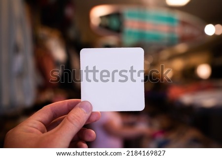 Hand holding a blank white square paper card with rounded corners. Business card, magnet, bumper sticker mock-up. Clothing retail store space background. 