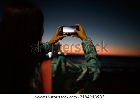 Trendy tourist taking photo on her phone of the ocean with beautiful sunset on the horizon at night outdoors. Influencer or vlogger taking picture of nature sea view to post or share on social media