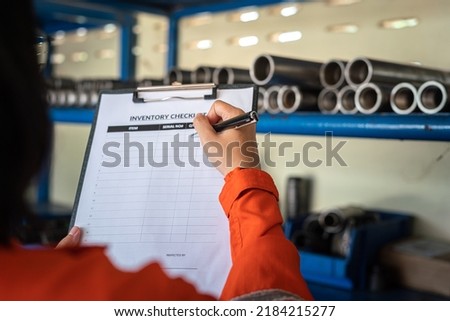 A worker is using pen to writing on inventory checklist form, with blurred background of storage shelf in the factory. Industrial working action photo. Selective focus.