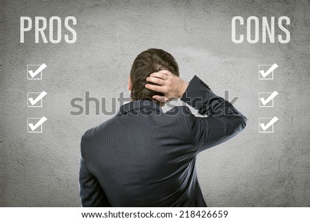 Young businessman selecting all pros and cons in front of grey wall. Portrait of financial manager comparing advantages and risks of investment deal.   Royalty-Free Stock Photo #218426659
