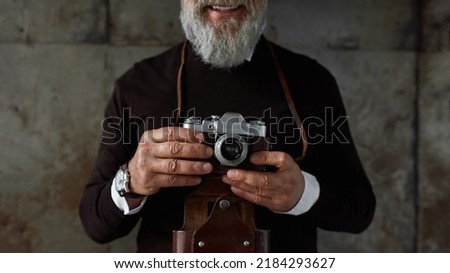 Obscure face of senior smiling man with old camera. Stylish bearded pensioner. Modern elderly male lifestyle. Entertainment and hobby. Isolated on rusty concrete background. Studio shoot