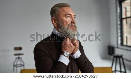 Cropped image of senior serious caucasian man correcting shirt collar in spacious apartment. Stylish bearded pensioner wearing casual clothes. Concept of modern elderly male lifestyle. Daytime
