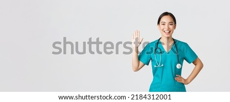 Covid-19, healthcare workers and preventing virus concept. Friendly-looking smiling asian female doctor, physician in scrubs waving hand to say hi, hello, greeting patient, nice to see you Royalty-Free Stock Photo #2184312001