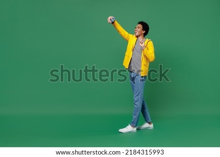 Full size young black curly man 20s wears yellow waterproof raincoat outerwear doing selfie shot on mobile cell phone post photo on social network isolated on plain green background studio portrait
