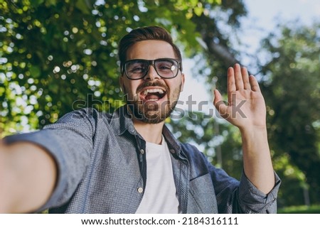 Close up young fun man 20s in blue shirt glasses do selfie shot on mobile cell phone waving hand rest relax in spring forest garden green city park outdoors on nature Urban lifestyle leisure concept