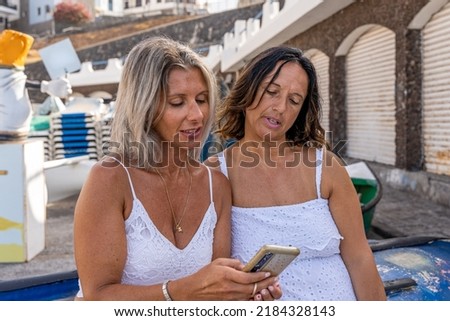 two beautiful middle aged friends chatting sitting on an upturned boat in a small harbor