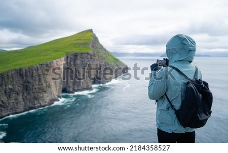 Back view of woman wearing winter clothes taking pictures Sorvagsvatn lake on Vágar Island. Tourist popular attraction destination in Faroe Islands. Young tourist woman taking photo with smart phone. 
