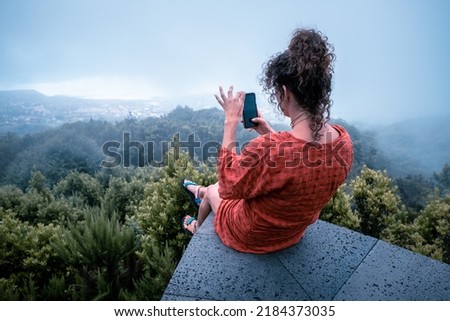 Young woman dressed in red sitting on the edge of a cliff taking a photo with her cell phone in a forest on a cloudy day with selective focus
