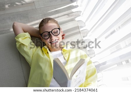 Young beautiful woman wearing yellow shirt reading a book at home. Brunette female lying on the textile couch in her lofty apartment and studying. Background, copy space, close up. Royalty-Free Stock Photo #2184374061