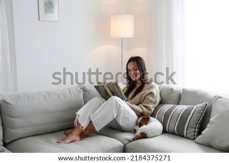 Young beautiful woman reading a book at home with her wire haired jack russell terrier pup. Brunette female and rough coated dog sitting on textile couch. White wall background, copy space, close up. Royalty-Free Stock Photo #2184375271