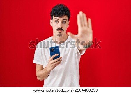 Hispanic man using smartphone over red background doing stop sing with palm of the hand. warning expression with negative and serious gesture on the face. 