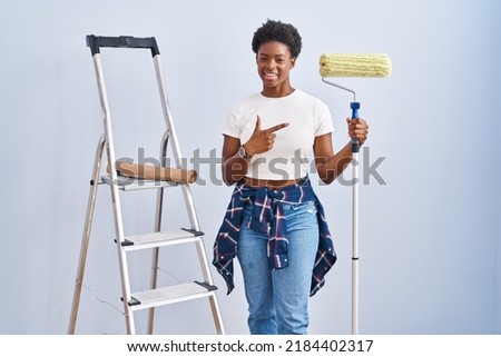 African american woman holding roller painter cheerful with a smile on face pointing with hand and finger up to the side with happy and natural expression 