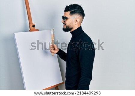Handsome man with beard holding brushes close to easel stand looking to side, relax profile pose with natural face and confident smile. 