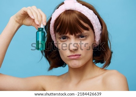 a sad, anxious woman stands on a blue background fastidiously holding a facial serum in her hand and looks into the camera expressing negative emotions. Horizontal photo