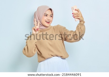 Cheerful young Asian muslim woman in brown sweater doing selfie shot on mobile phone, make peace sign isolated over white background. People religious lifestyle concept
