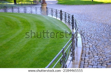watering lawn automatic irrigation with pull-out sprinklers fresh green color black plastic nozzles extend and rotate in a circular rotation water granite cobblestone tiles and curb, fence, railings Royalty-Free Stock Photo #2184465555