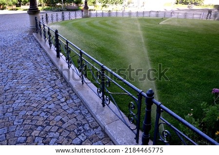 watering lawn automatic irrigation with pull-out sprinklers fresh green color black plastic nozzles extend and rotate in a circular rotation water granite cobblestone tiles and curb, fence, railings Royalty-Free Stock Photo #2184465775