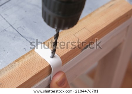 A man drills holes in an iron hook with a drill to hang a wooden screen on a battery.