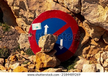 No parking sign leaning against a rock and covered with stones.