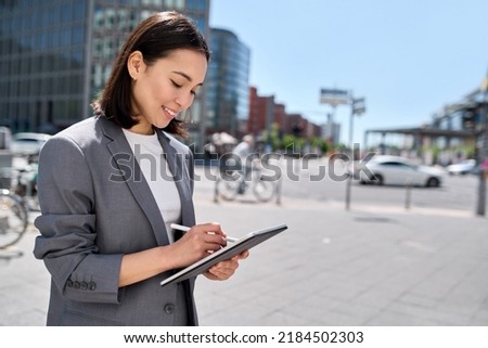 Young happy Asian business woman executive or saleswoman holding digital tablet using software applications standing on the street in big city enjoying smart technology data concept.