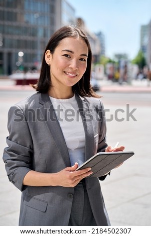 Young happy Asian business woman entrepreneur wearing suit holding digital tablet standing on big city busy street using smart business software tech for online work on pad computer outdoor Royalty-Free Stock Photo #2184502307