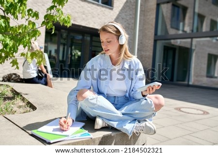 Pretty girl university student wearing headphones using smartphone app sitting on stairs outdoors in campus online learning, remote studying class, watching educational webinar web distance course. Royalty-Free Stock Photo #2184502321