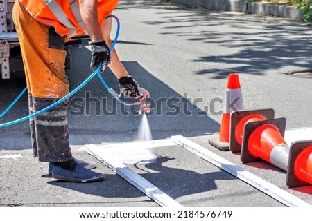 An airbrush in the hands of a road worker applies white road markings to a pedestrian crossing using a wooden template and orange traffic cones. Copy space.