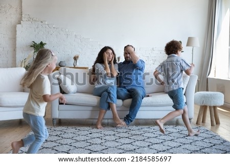 Two active sibling kids running around tired parents, playing active games. Mom and dad exhausted with watching children, feeling headache, stress. Upbringing problems, parenthood, family concept Royalty-Free Stock Photo #2184585697