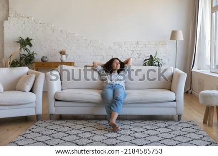 Happy thoughtful young Latin woman resting on comfortable couch at home, looking away, smiling, dreaming, thinking, relaxing, enjoying leisure time, breathing fresh air Royalty-Free Stock Photo #2184585753