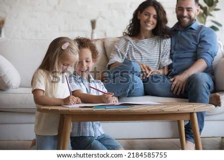 Happy satisfied young mother and father relaxing on sofa, watching two cute little sibling children drawing in colored pencils, playing in living room, smiling, enjoying home leisure, family