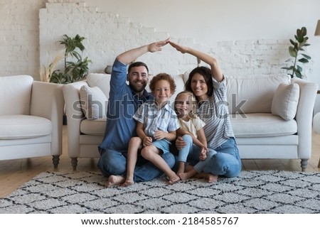 Happy millennial family couple and two little children hugging under hand home roof, sitting on heating floor in cozy living room, smiling at camera. Real estate, house buying, insurance concept
