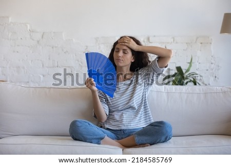 Tired annoyed Latin woman waving paper handheld fan for cooling air, suffering from hot summer weather, heat attack symptoms, headache, migraine, touching head with closed eyes Royalty-Free Stock Photo #2184585769