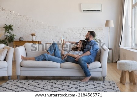 Happy millennial couple of homeowners enjoying cool conditioned air, resting on couch together, using remote control for AC, cooling domestic equipment start. Home appliance concept Royalty-Free Stock Photo #2184585771