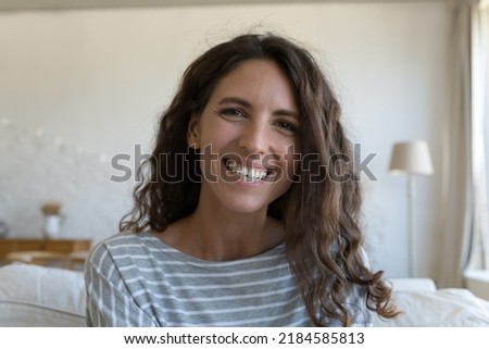 Happy pretty young adult Latin woman home head shot portrait. Cheerful female model sitting on couch, looking at camera, posing with toothy smile, laughing, speaking on video call