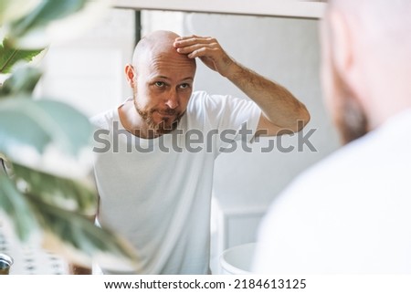 Young adult bearded man looking in mirror in bathroom touching head worried about about hair loss Royalty-Free Stock Photo #2184613125