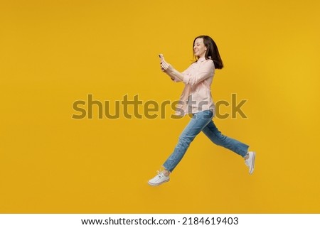 Full body side view young fun woman she 30s in striped shirt white t-shirt jump high run fast hurry up hold use mobile cell phone isolated on plain yellow background studio. People lifestyle concept.