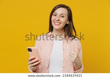 Young satisfied fun happy woman she 30s wears striped shirt white t-shirt hold in hand use mobile cell phone chatting onlie browsing internet show thumb up isolated on plain yellow background studio. Royalty-Free Stock Photo #2184619421