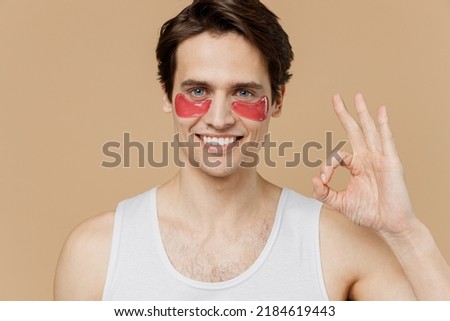 Attractive fun young man he 20s perfect skin in undershirt wearing gold patch under eye show ok okay isolated on plain pastel beige background studio. Skin care healthcare cosmetic procedures concept