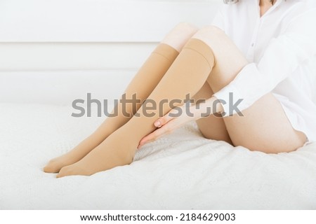 Beautiful long female legs in stockings. Girl putting on stockings at home in a white room. Beige knee socks. Varicose veins prevention. Woman body in underwear.