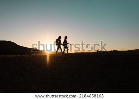 silhouette of two girls with backpacks while traveling in the mountains at sunset. two people walk along the mountain range at sunset. travel with freedom. Royalty-Free Stock Photo #2184658163