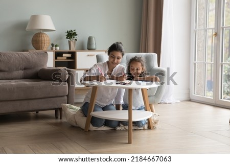 Engaged young Indian mother and happy little daughter kid drawing in pencils in living room, scratching doodles in paper album, sitting at small table, chatting, laughing. Family home leisure