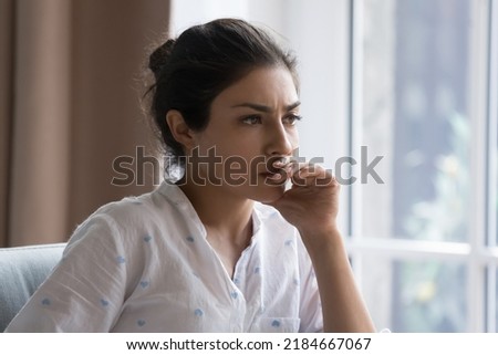 Serious thoughtful beautiful Indian woman sitting on couch indoors, touching chin, looking away, thinking over bad concerning news, problem, feeling worried, anxious, stressed, making decision Royalty-Free Stock Photo #2184667067
