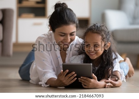 Happy Indian little kid and mom using digital tablet, resting on heating floor at home, smiling, laughing. Mother and child enjoying leisure, watching movie, reading book online