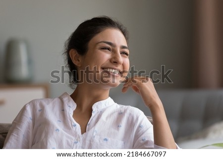Happy beautiful young Indian woman looking at window with toothy smile, thinking of future vision, good news, dreaming, touching chin, facial skin, laughing. Head shot portrait