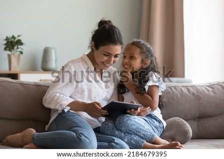 Happy Indian little daughter kid talking to mom, using digital tablet, sitting on sofa at home, smiling, laughing. Mother and child enjoying leisure, playing game on digital gadget Royalty-Free Stock Photo #2184679395